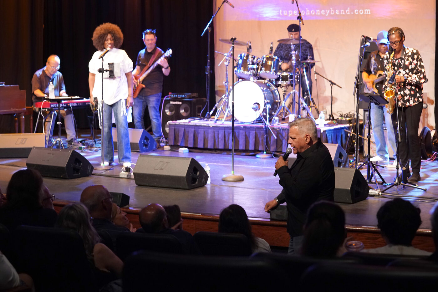 Full band performing on stage with tupelohoneyband.com banner